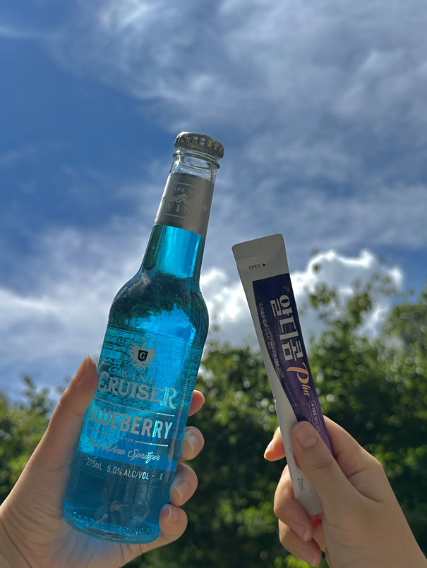 Hands holding a blue alcoholic drink and a stick of Aldicom hangover relief under a bright sky—showcasing smart drinking with Aldicom for next-day wellness.
