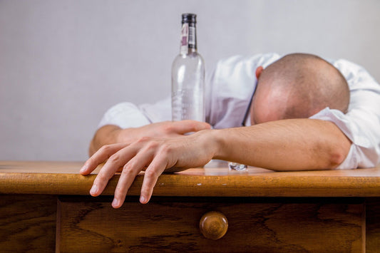 Man passed out with his head on the table next to an empty vodka bottle—depicting severe hangover fatigue and the perfect use case for Aldicom’s recovery supplement