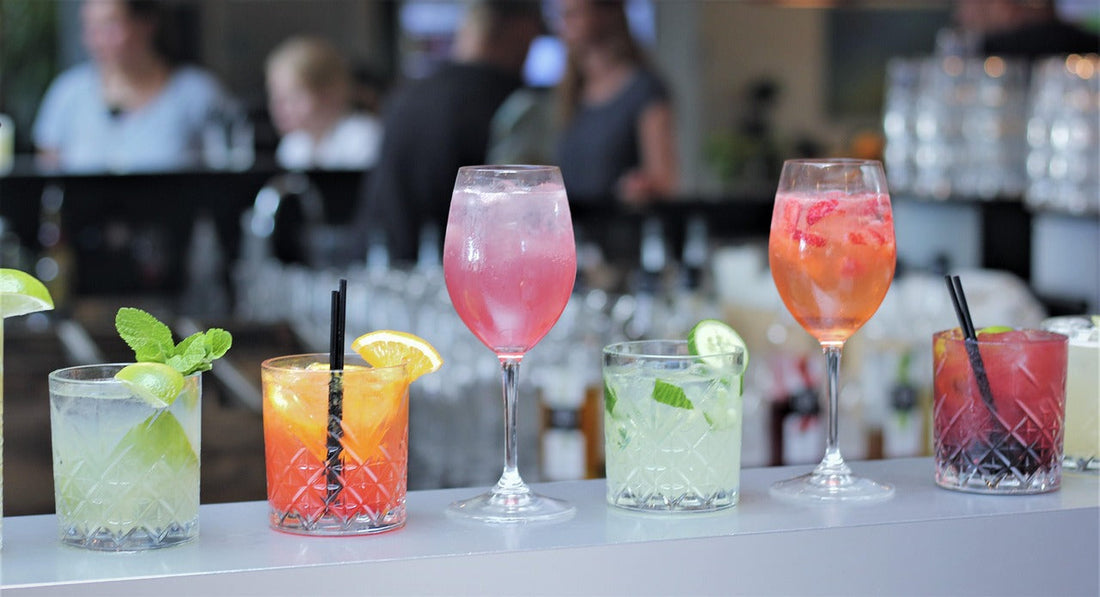 Colorful row of cocktails on a bar counter with people in the background—perfect setting to enjoy drinks responsibly with Aldicom hangover prevention on hand