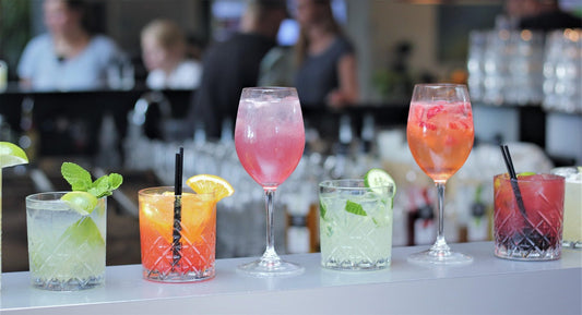 Colorful row of cocktails on a bar counter with people in the background—perfect setting to enjoy drinks responsibly with Aldicom hangover prevention on hand
