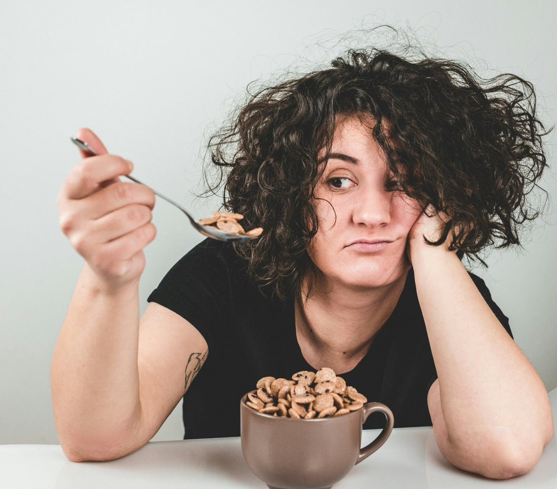 Tired woman with messy hair holding a spoonful of cereal, looking exhausted at the breakfast table—representing morning fatigue and the need for Aldicom hangover relief